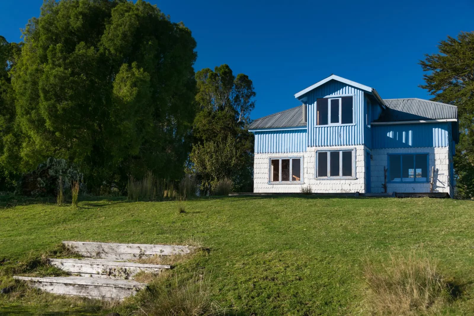 Casa de campo de estilo sureño, azul y blanca, rodeada de árboles nativos bajo un cielo despejado
